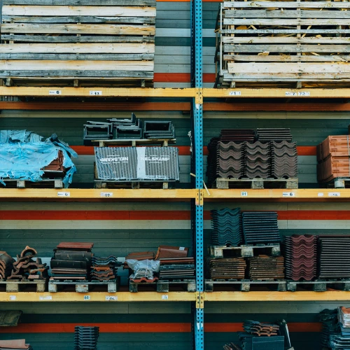 Shelving in a warehouse stocked with stacked roofing tiles, metal parts, and construction materials, organized across multiple pallet racks in an industrial setting.