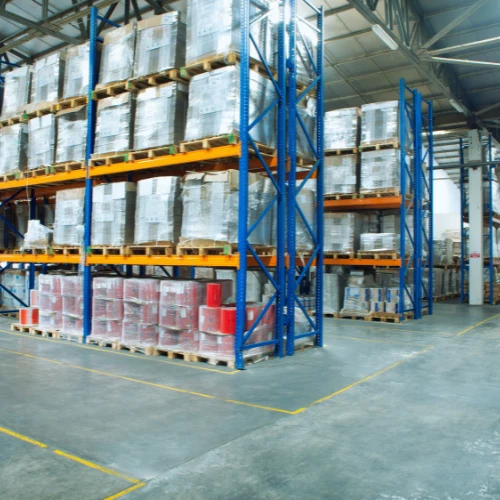 Wide-angle view of a warehouse with tall blue and yellow pallet racking, stacked with shrink-wrapped goods and bulk inventory, organized on polished concrete floors.